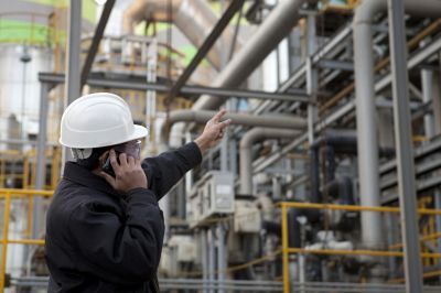 Homem com capacete de segurança se comunica via celular e aponta em direção a tubulações em ambiente industrial